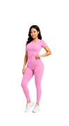 A woman modeling a bright pink short-sleeve athletic jumpsuit with white sneakers, standing against a light gray studio background.