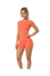 A woman modeling a coral orange short-sleeve athletic romper with matching fitted shorts against a plain white background.