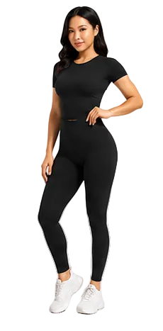 A woman modeling a black short-sleeve crop top and matching high-waisted leggings set, paired with white sneakers, against a light gray studio background.