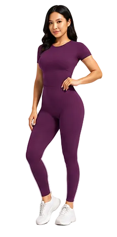 A woman modeling a short-sleeve, high-waisted, form-fitting athletic jumpsuit in a solid deep purple color, paired with white sneakers, against a light gray studio background.