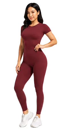 A woman modeling a short-sleeve, high-waisted burgundy athletic jumpsuit with white sneakers, set against a plain light gray studio background.