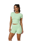 A woman modeling a light mint green two-piece activewear set consisting of a short-sleeve ribbed crop top and matching high-waisted shorts, set against a plain white background.